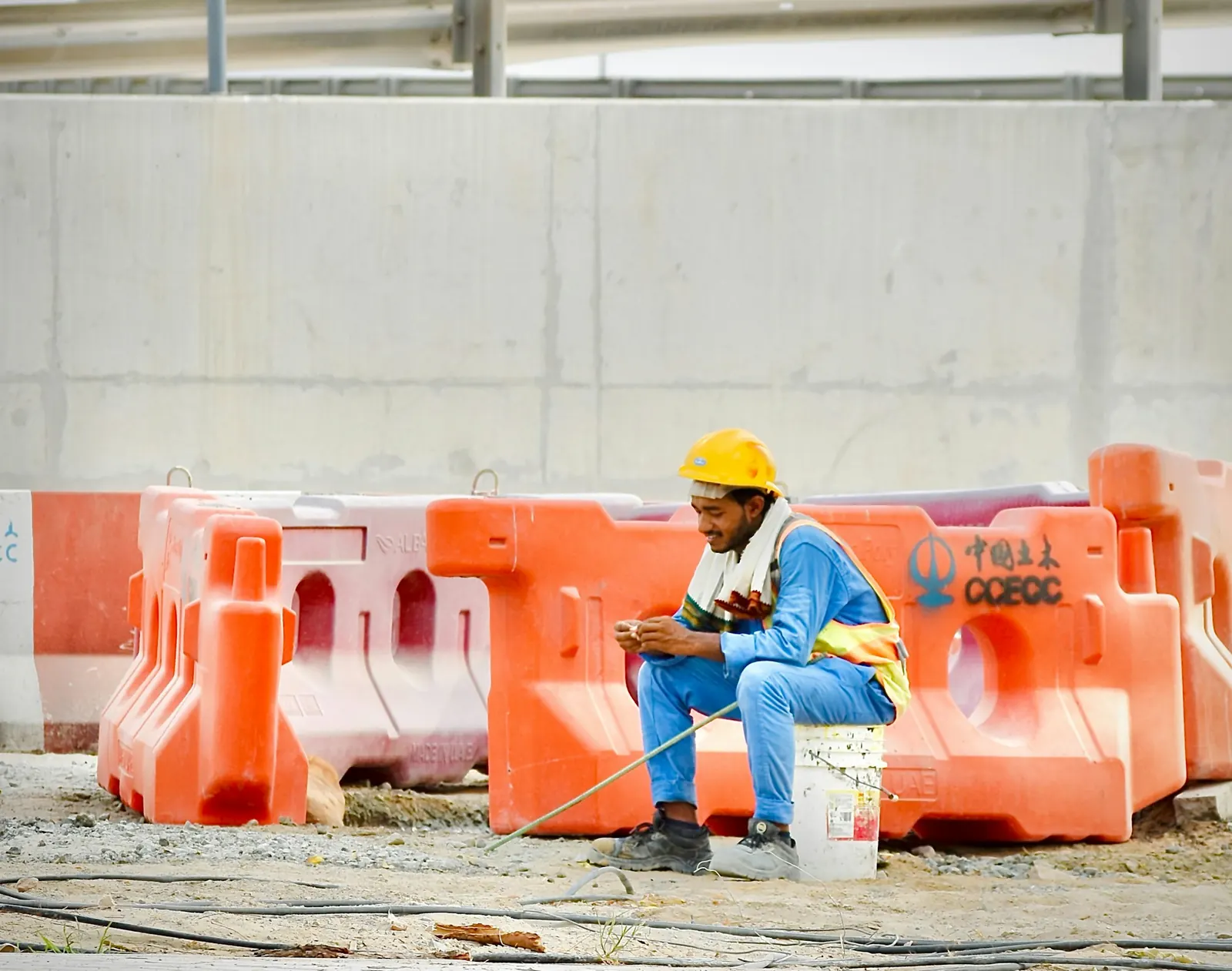 A builder in a hard hat sitting on an upturned bucket on a building site, checking his phone