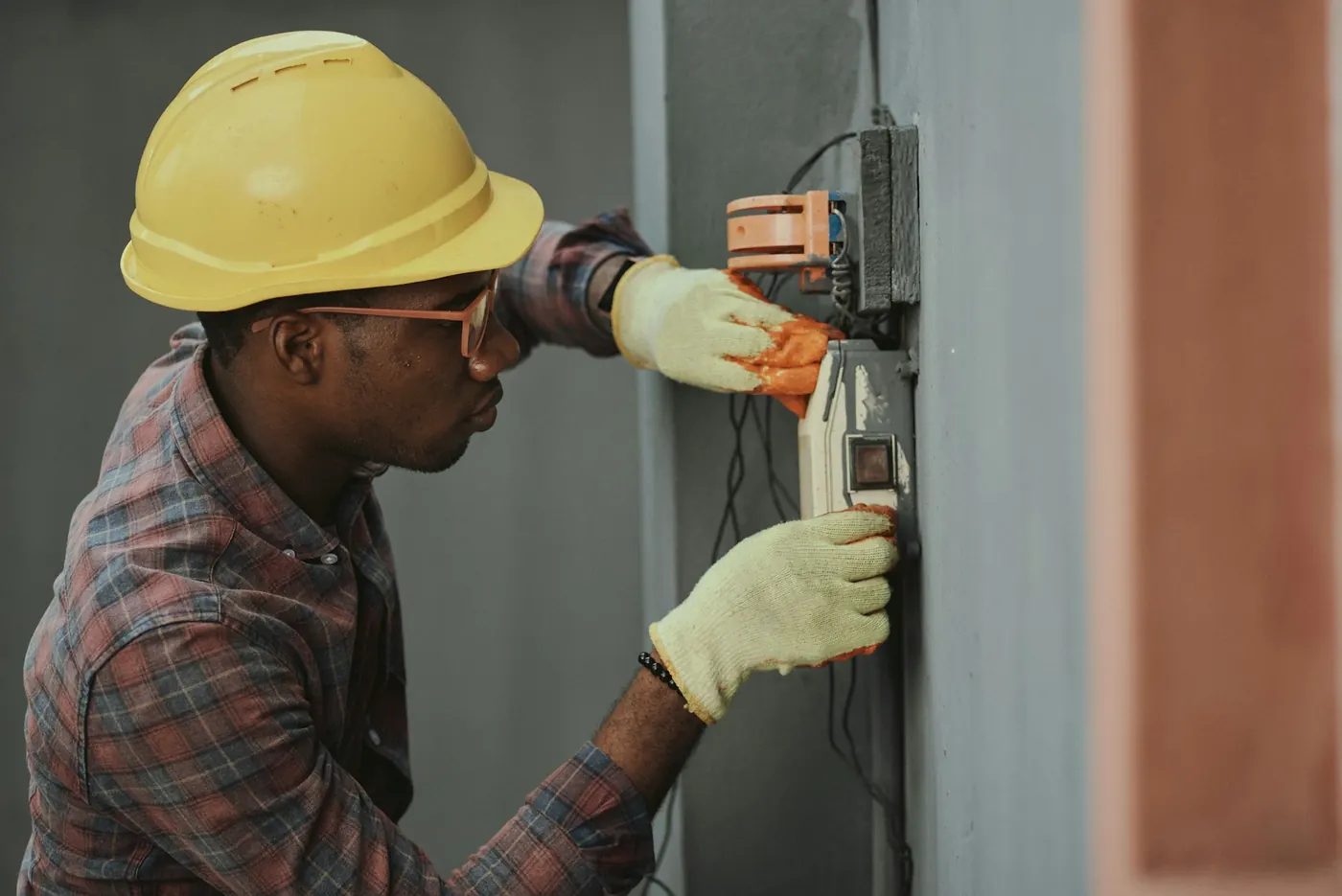 An electrician in a yellow hard hat and safety glasses testing wiring in a wall-mounted panel